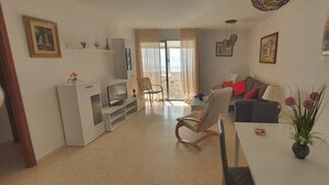 Ein Wohnzimmer mit Fernseher, Sofa, Esstisch und Stühlen. Ein Fenster mit Vorhängen, ein Gemälde an der Wand und eine Vase mit Blumen auf dem Tisch.