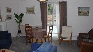 Un salon avec un canapé bleu, une table à manger avec des chaises, une plante en pot et une vue à travers une porte vitrée.