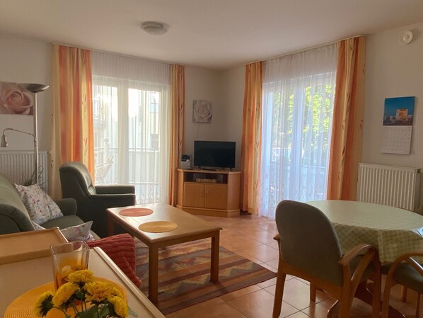 A cozy living room with a sofa, a dining table, a TV, and a view of the outdoors through large windows.