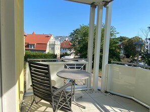 A balcony with a metal chair and a small round table, overlooking a street with buildings and trees.