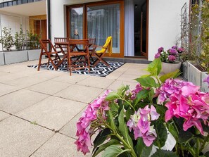 Eine Terrasse mit Tisch und Stühlen, ein gelber Stuhl, ein gemustertes Teppich und Blumentöpfe mit rosa Blüten.