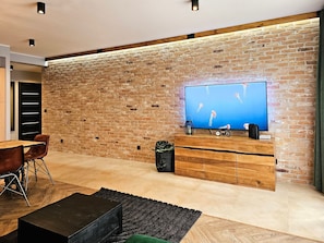Een moderne woonkamer met een bakstenen muur, een flatscreen-tv, houten kasten, een eethoek met stoelen en een zwarte salontafel.