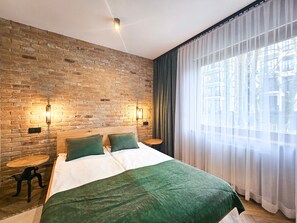 Une chambre avec un mur en briques, deux tables de chevet, un lit avec une literie verte et une grande fenêtre ornée de rideaux légers.