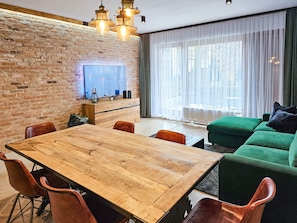 Un salon moderne avec un mur en briques, une table à manger en bois, un canapé vert et une grande fenêtre ornée de rideaux légers.