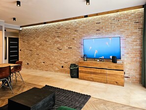 A modern living room with a brick wall, a flat-screen TV, wooden cabinets, a dining area with chairs, and a black coffee table.
