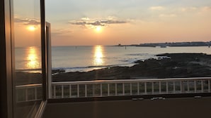 Une vue sur le coucher de soleil en bord de mer, prise depuis un balcon doté d’une balustrade, avec une côte rocheuse et une silhouette de ville lointaine.