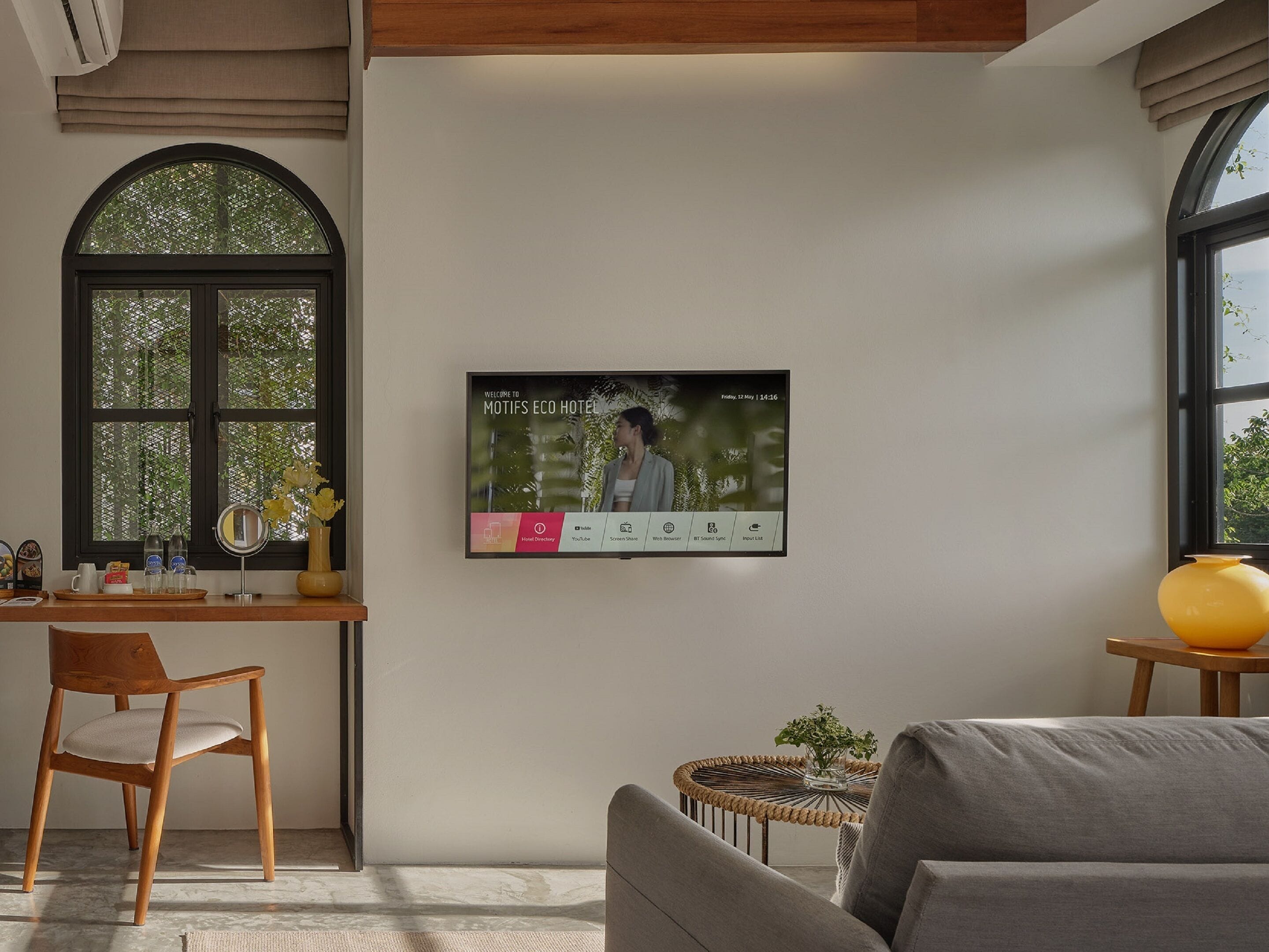 A modern hotel room with a large arched window, a desk with a vase of flowers, a comfortable sofa, and a wall-mounted TV displaying a welcome message.