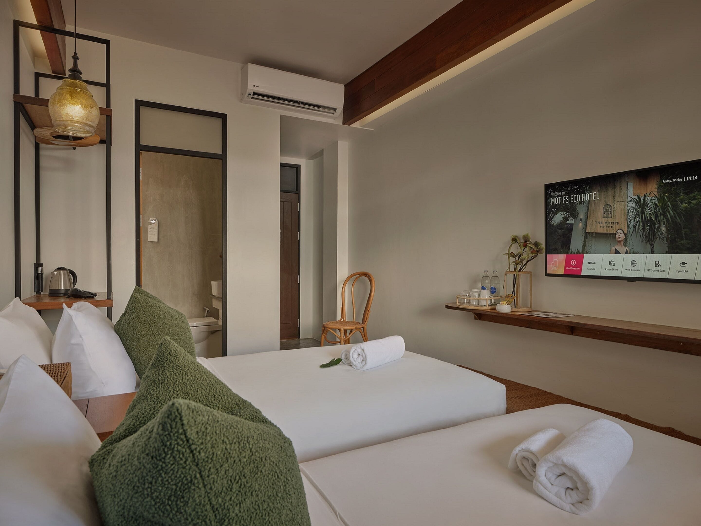 Una habitación de hotel con cama, almohadas, una silla, un televisor y un estante con artículos de aseo.