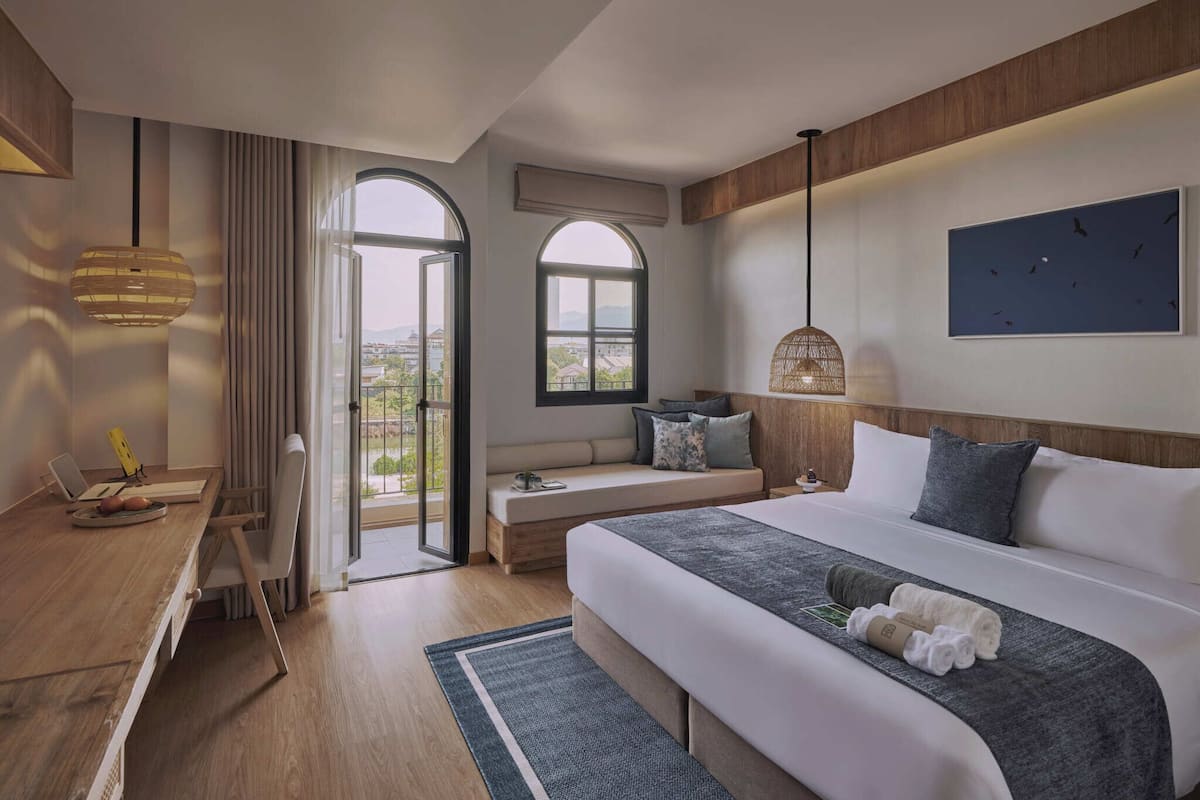 Une chambre d’hôtel avec un grand lit, un bureau avec un ordinateur portable, une vue à travers une porte vitrée et un luminaire suspendu.