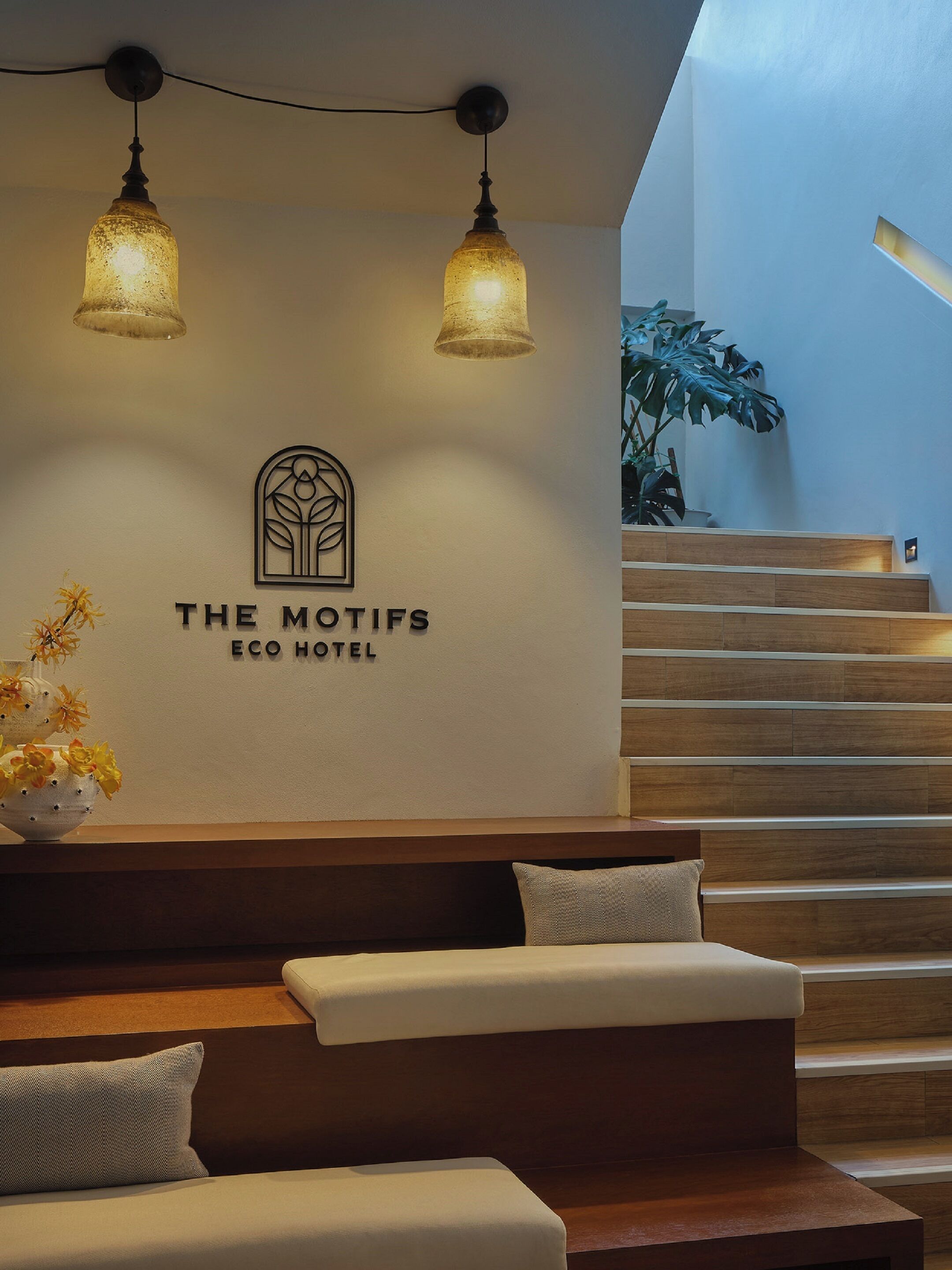 Ein Empfangsbereich mit einem Schild für "The Motifs Eco Hotel", einer hölzernen Sitzbank mit Kissen und einer Treppe.