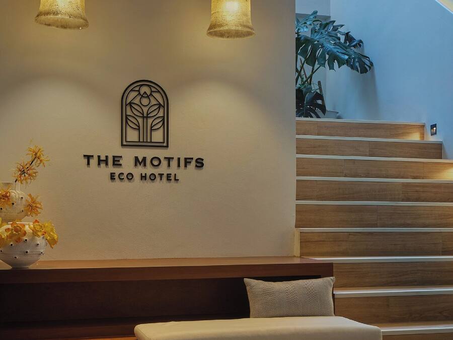 Ein Empfangsbereich mit einem Schild für "The Motifs Eco Hotel", einer hölzernen Sitzbank mit Kissen und einer Treppe.