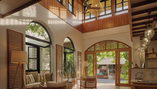 A spacious living area with wooden flooring, a high ceiling, and large windows allowing natural light. There are multiple seating areas with cushions and a coffee table. - The Motifs Eco Hotel (Chanthaburi)