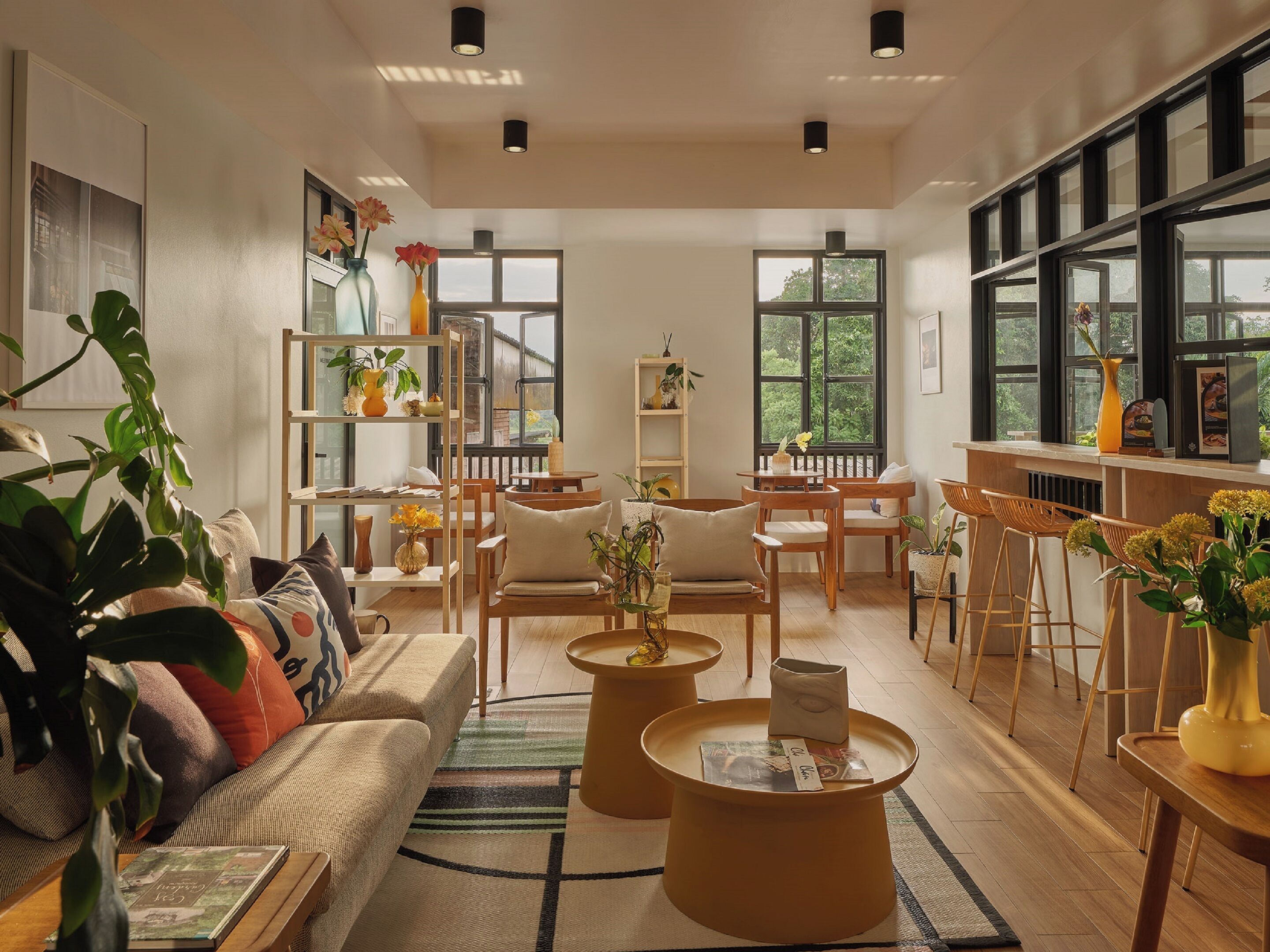 A spacious living room with a sofa, coffee table, and bar stools. There are shelves with plants and a large window providing natural light.