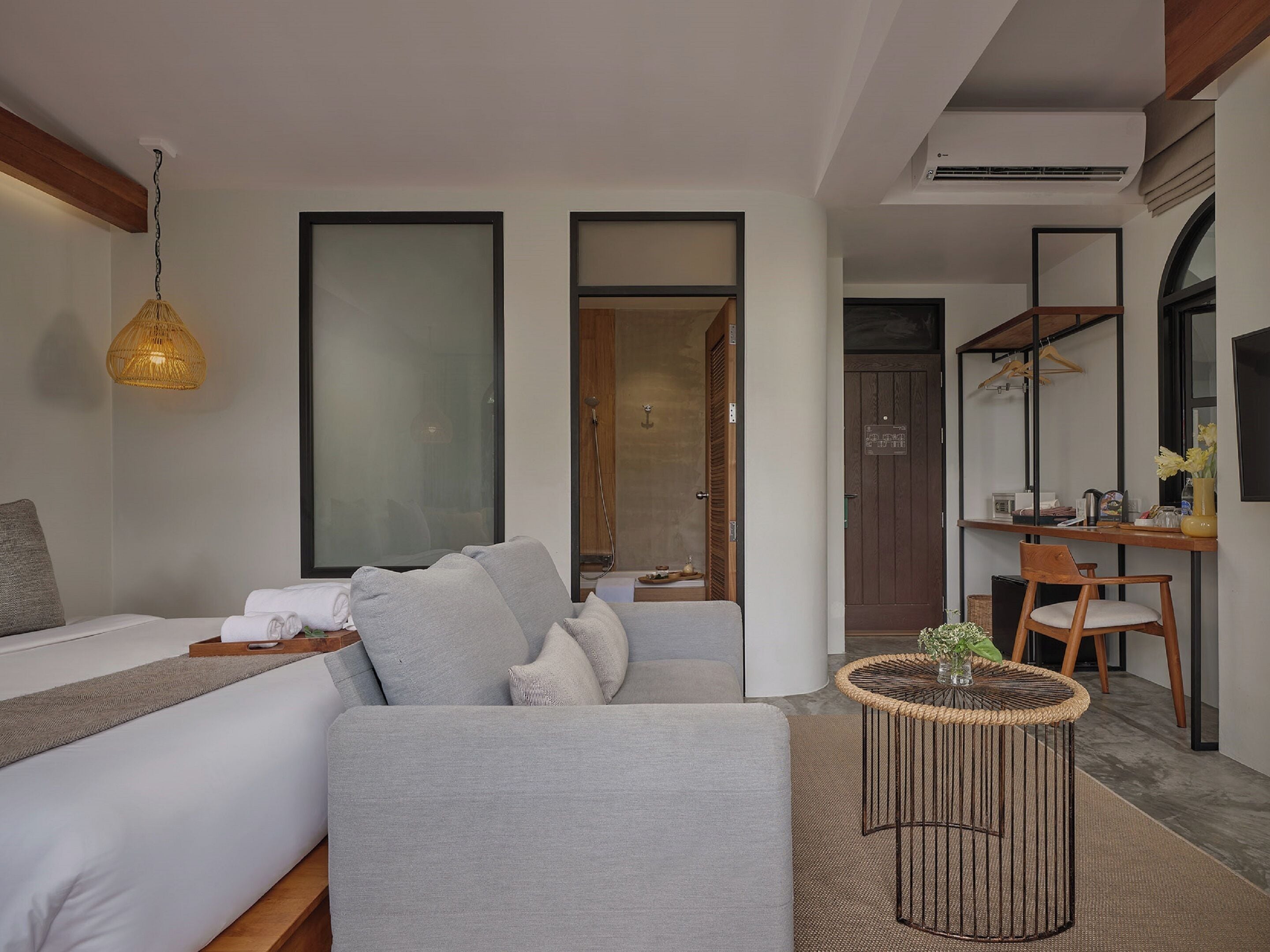 A modern hotel room with a grey sofa, a small round table with a plant, a hanging light fixture, and a wooden desk with a mirror and a hanging coat rack.