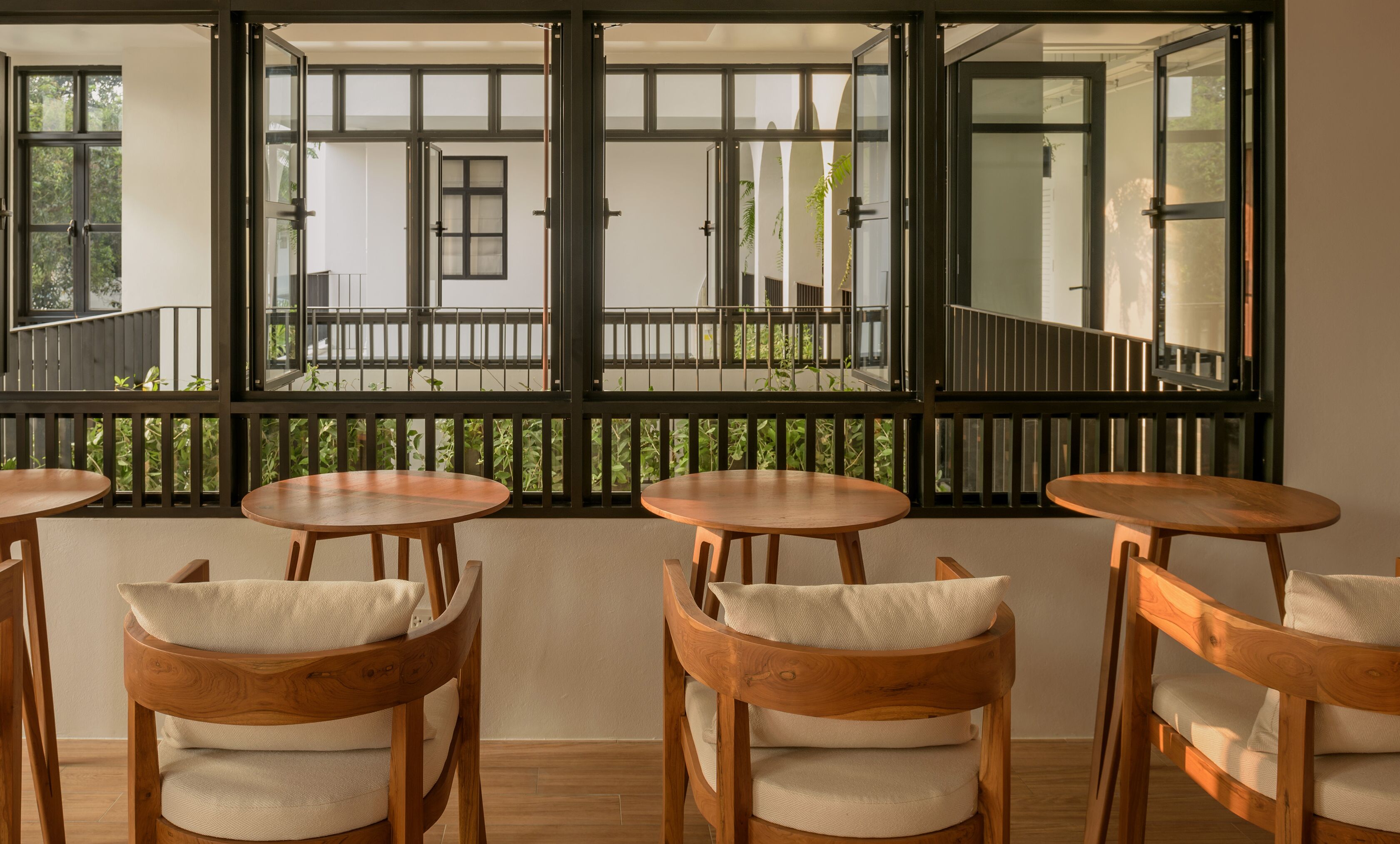 Una habitación con cuatro mesas y sillas de madera, ventanas amplias que dan a un balcón y follaje exterior.