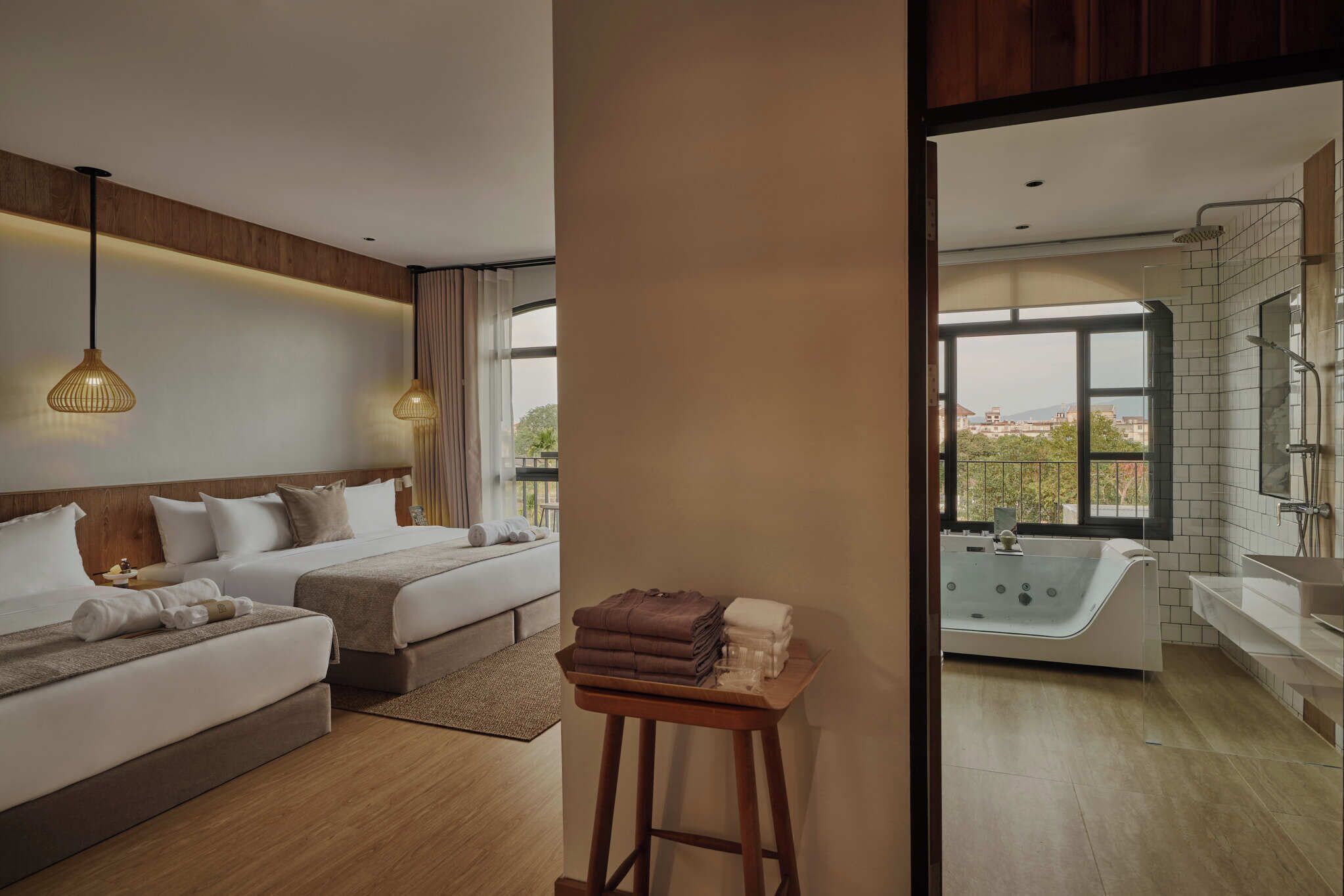 Habitación de hotel con dos camas, una zona de estar, una mesita con toallas, una bañera y un ventanal con vistas a la ciudad.