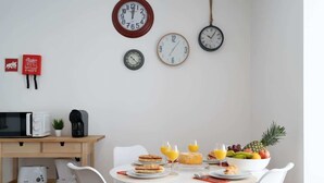 Ein Frühstückstisch mit weißen Stühlen und einer Auswahl an Speisen, darunter Toast, Obst und Saft. An der Wand sind Uhren angebracht und auf der Arbeitsfläche steht eine Mikrowelle.