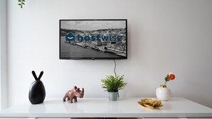 Ein weißer Schreibtisch mit einem Fernseher, der eine Stadtansicht zeigt, eine schwarze Vase, eine rosa Elefantenfigur, eine Topfpflanze und eine weiße Vase mit einer Blume.