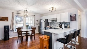 Cozinha moderna com armários brancos, uma ilha central com banquetas, uma mesa de jantar com cadeiras e uma geladeira.
