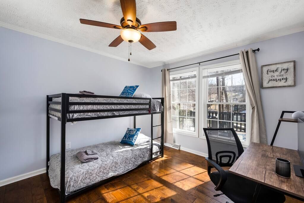 A room with a bunk bed, a ceiling fan, a window with curtains, a desk with a chair, and a motivational quote on the wall.