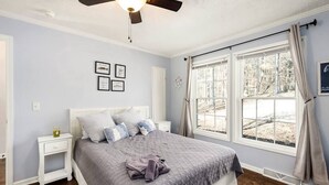 Quarto com cama, mesa de cabeceira, ventoinha no teto e janela com cortinas.