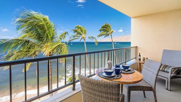A balcony with wicker furniture, a table set with a coffee pot, cups, and saucers, overlooking a beach with palm trees and the ocean. - Sugar Beach PH21 - Direct Oceanfront 1 bedroom (Kihei)