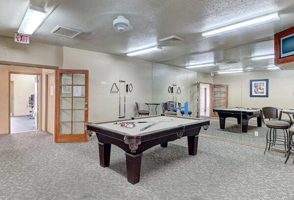 Une table de billard dans une pièce dont le sol est recouvert d’une moquette, avec une porte dotée d’un panneau en verre et un panneau indiquant la sortie.