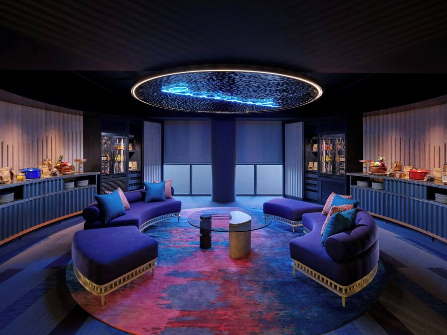 Ein moderner Lounge-Bereich mit violetten Sitzgelegenheiten, einem zentralen Couchtisch und einer Neon-Deckenleuchte.