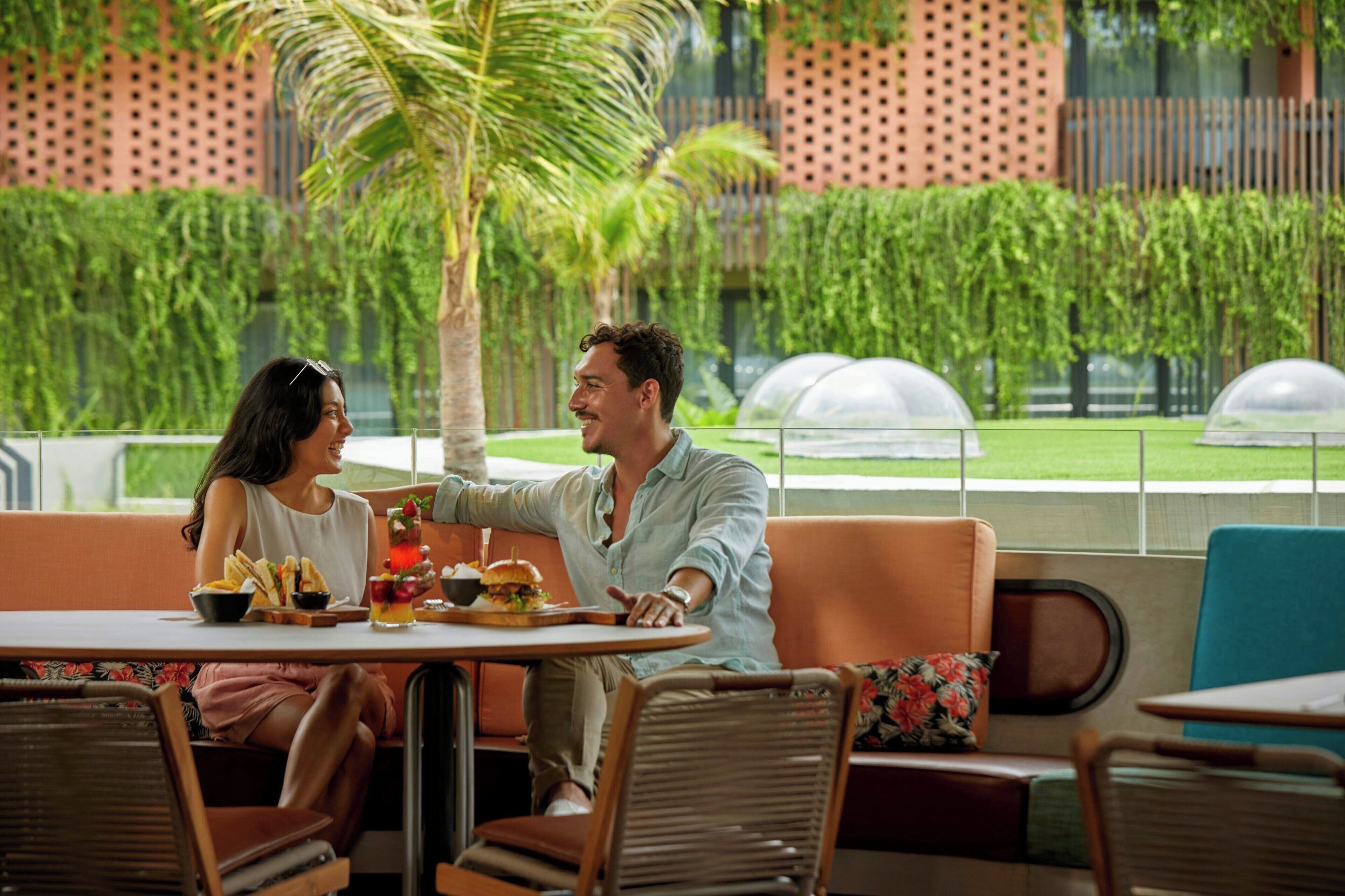 Una pareja cenando en un café al aire libre con vistas a un jardín exuberante y un edificio moderno al fondo.