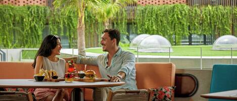 A couple dining at an outdoor cafe with a view of a lush garden and a modern building in the background.