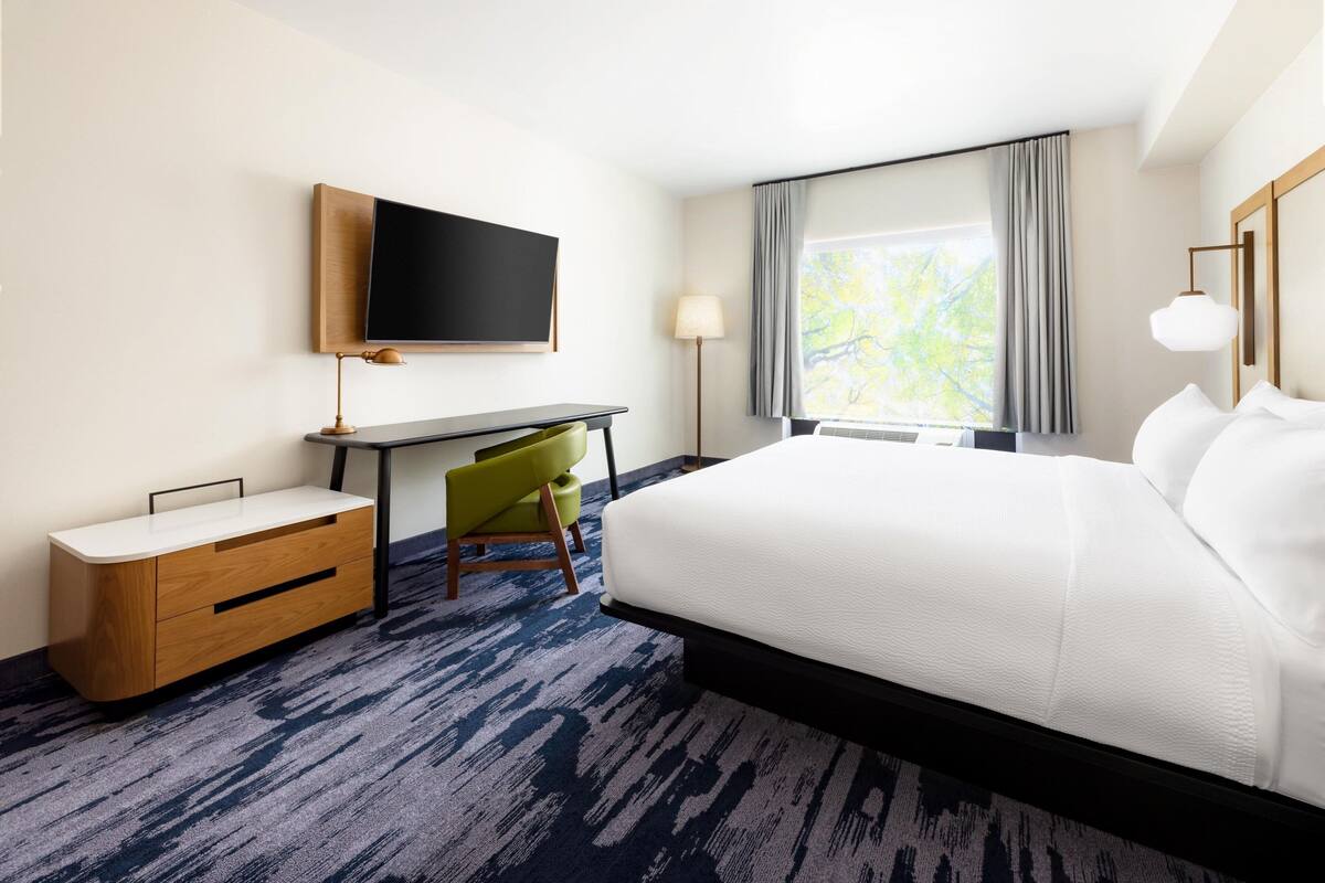 Ein Hotelzimmer mit einem großen Bett, einem Schreibtisch, einem Sessel, einem Fernseher und einem Fenster mit Vorhängen.