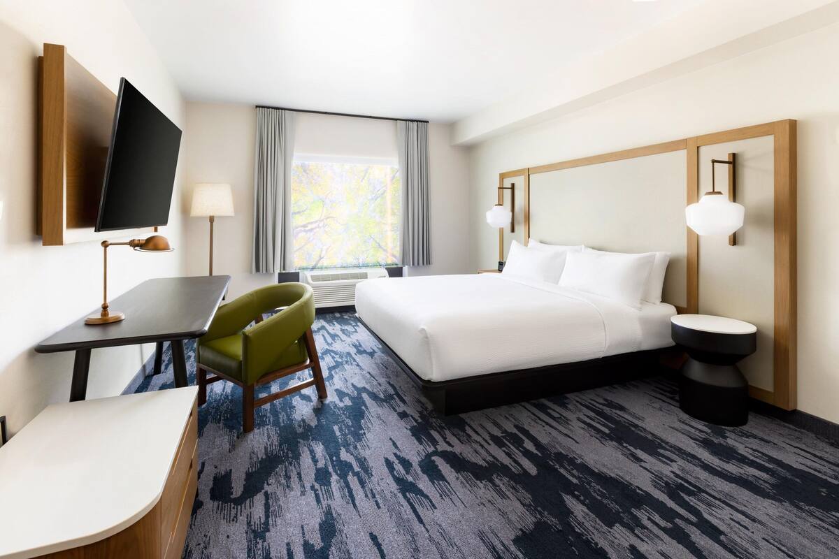 Ein Hotelzimmer mit Bett, Schreibtisch, Stuhl, Fernseher und einem Fenster mit Vorhängen.