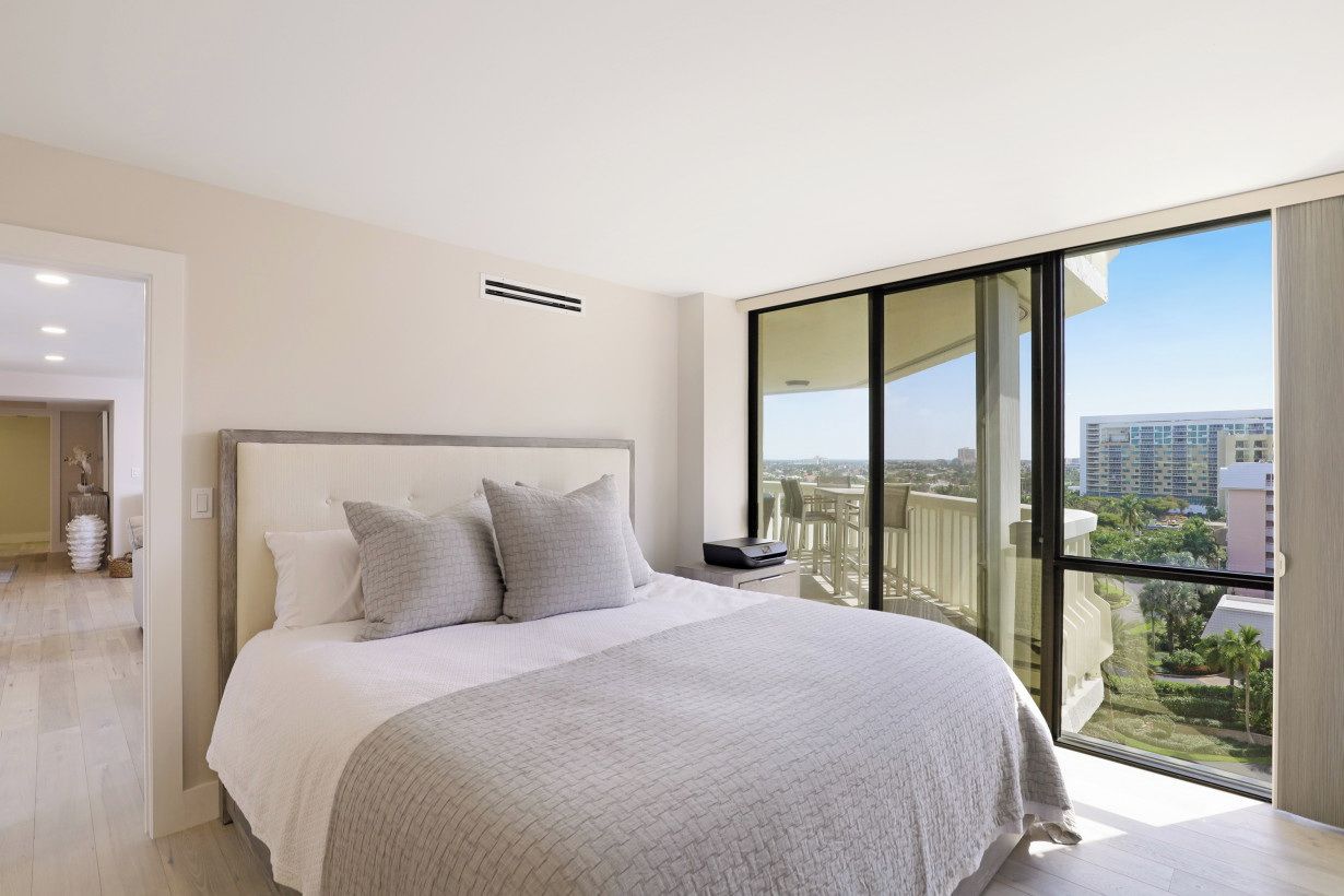 A modern bedroom with a large bed, a bedside table, and a view of a cityscape through floor-to-ceiling windows.