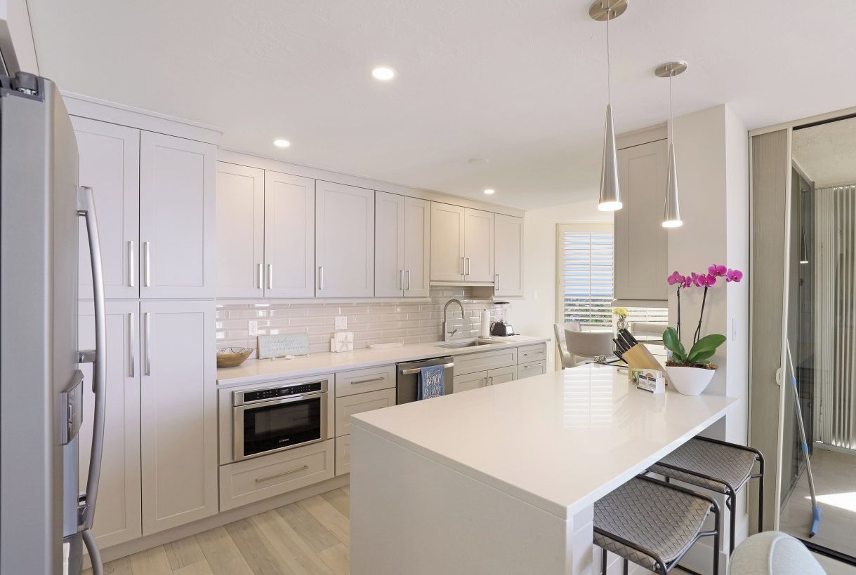 A modern kitchen with white cabinets, stainless steel appliances, a large island, pendant lights, and a potted plant.