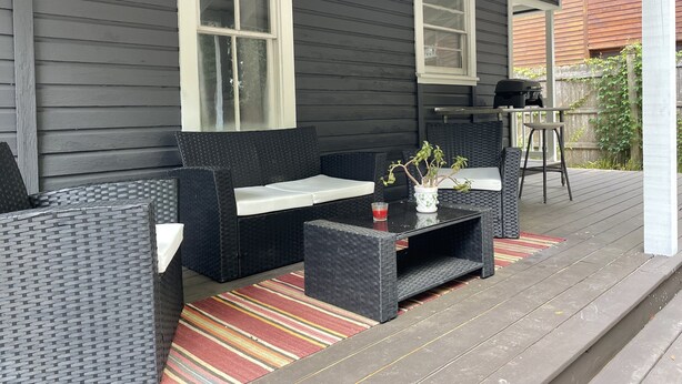 Een overdekte veranda met donkere bekleding, twee ramen, rieten meubels, een klein tafeltje en een gestreept tapijt.