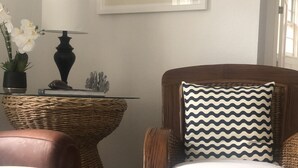 Een woonkamer met een rieten fauteuil, een glazen salontafel, een lamp en een ingelijst schilderij aan de muur.