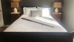 Een net opgemaakt bed met witte lakens, een houten knuppel en gestreepte kussens. Twee bedlampen staan op nachtkastjes.