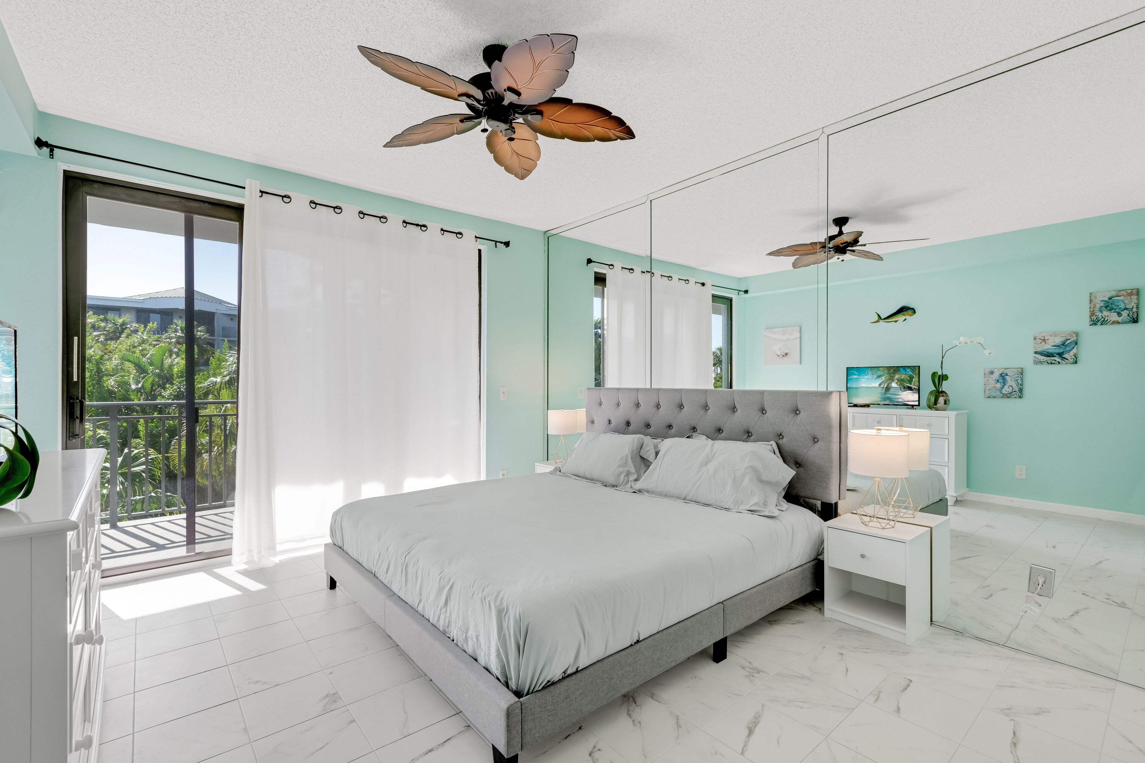 Une chambre moderne avec un grand lit, deux tables de chevet, un ventilateur de plafond et un balcon offrant une vue sur la verdure.