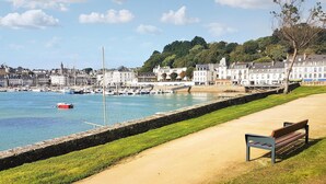 Une promenade en bord de mer avec un banc, un muret en pierre et un port de plaisance avec des bateaux.