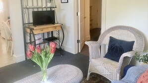Ett vardagsrum med en tv på ett träställ, en flätad fåtölj med en kudde, en vas med röda blommor och en dörr som leder till ett annat rum.