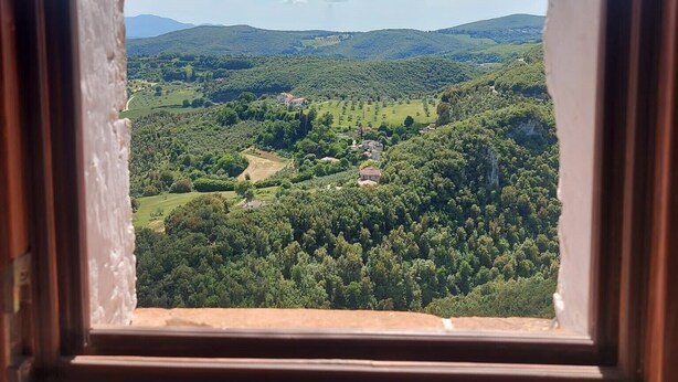 Vista a través d'una finestra de fusta, sobre un paisatge verd amb turons i edificis dispersos.
