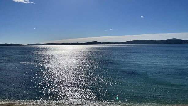 Vista clara de um mar calmo, com o sol refletindo na água, uma costa distante e céu limpo.