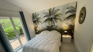 Quarto com cama grande, porta de vidro que desliza para um jardim e um mural na parede com palmeiras.