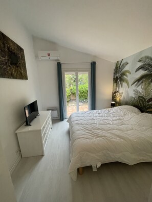 Une chambre avec un lit, un téléviseur posé sur une commode, une porte-fenêtre coulissante donnant sur un jardin et un climatiseur fixé au mur.