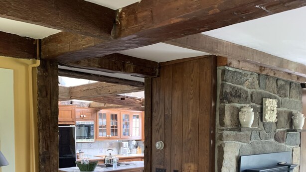 Un intérieur en bois avec une cheminée en pierre, des poutres apparentes et une cuisine à l’arrière-plan.