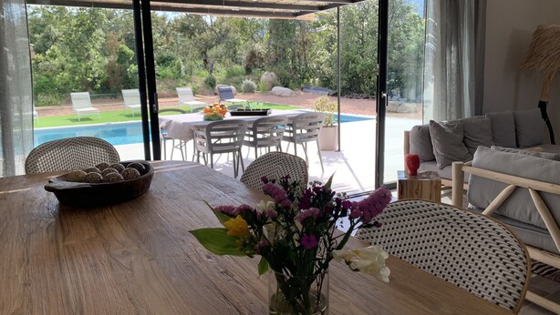 Ein Essbereich mit Holztisch, Stühlen und einem Blumenstrauß. Draußen befindet sich ein Pool und ein Garten mit Stühlen und einem Tisch.