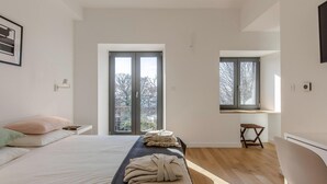 Ein modernes Schlafzimmer mit einem großen Bett, einer Bank mit Handtüchern und einem Fenster mit Blick auf Bäume.