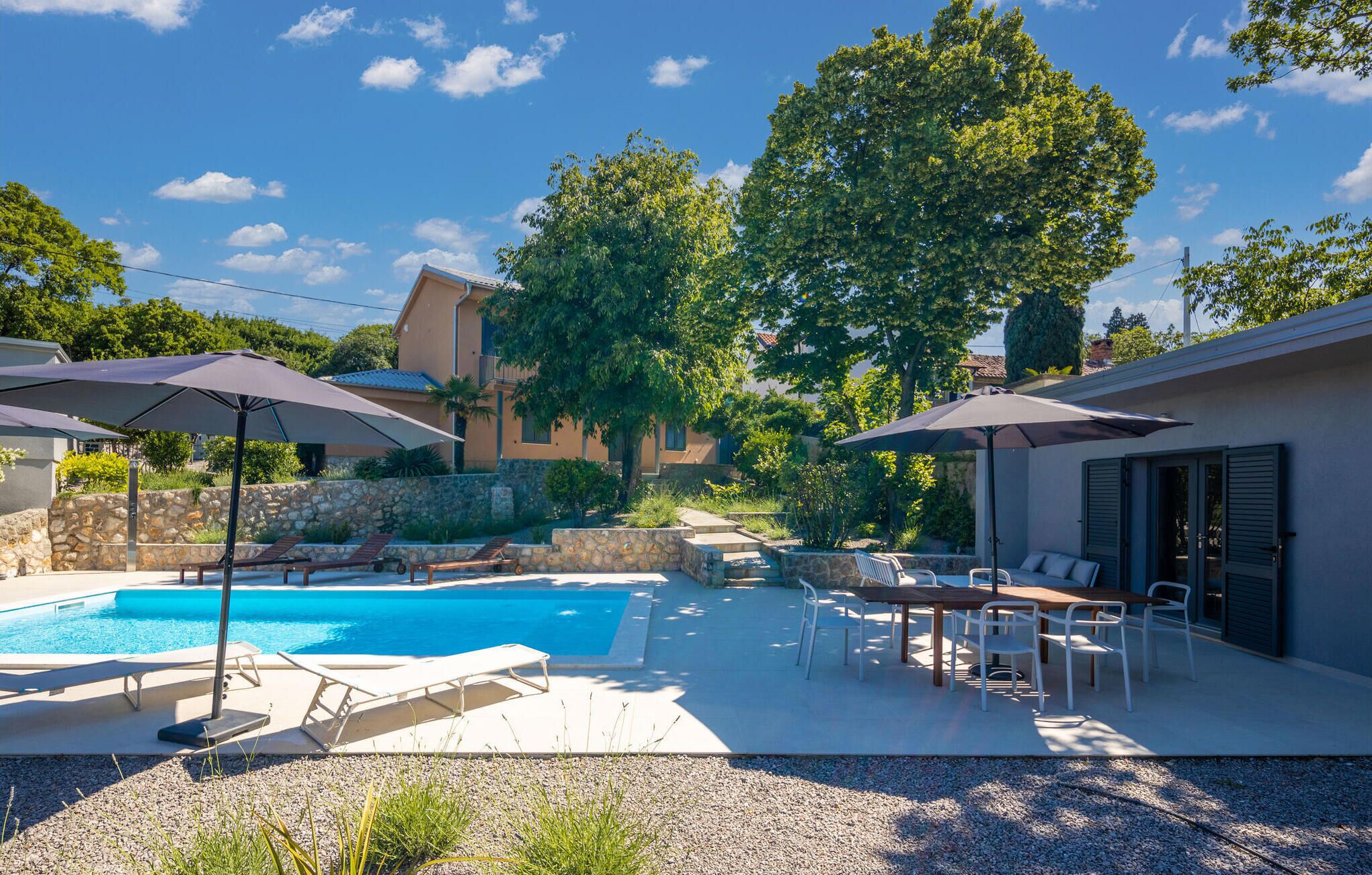 A pool area with lounge chairs and umbrellas, a stone wall, and a building with a balcony.