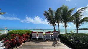 A wooden deck with white lounge chairs and red cushions, leading to a beach with clear turquoise waters and palm trees. - Exuma Sol - Stunning Views from this Oceanfront Beach Cottage (George Town)