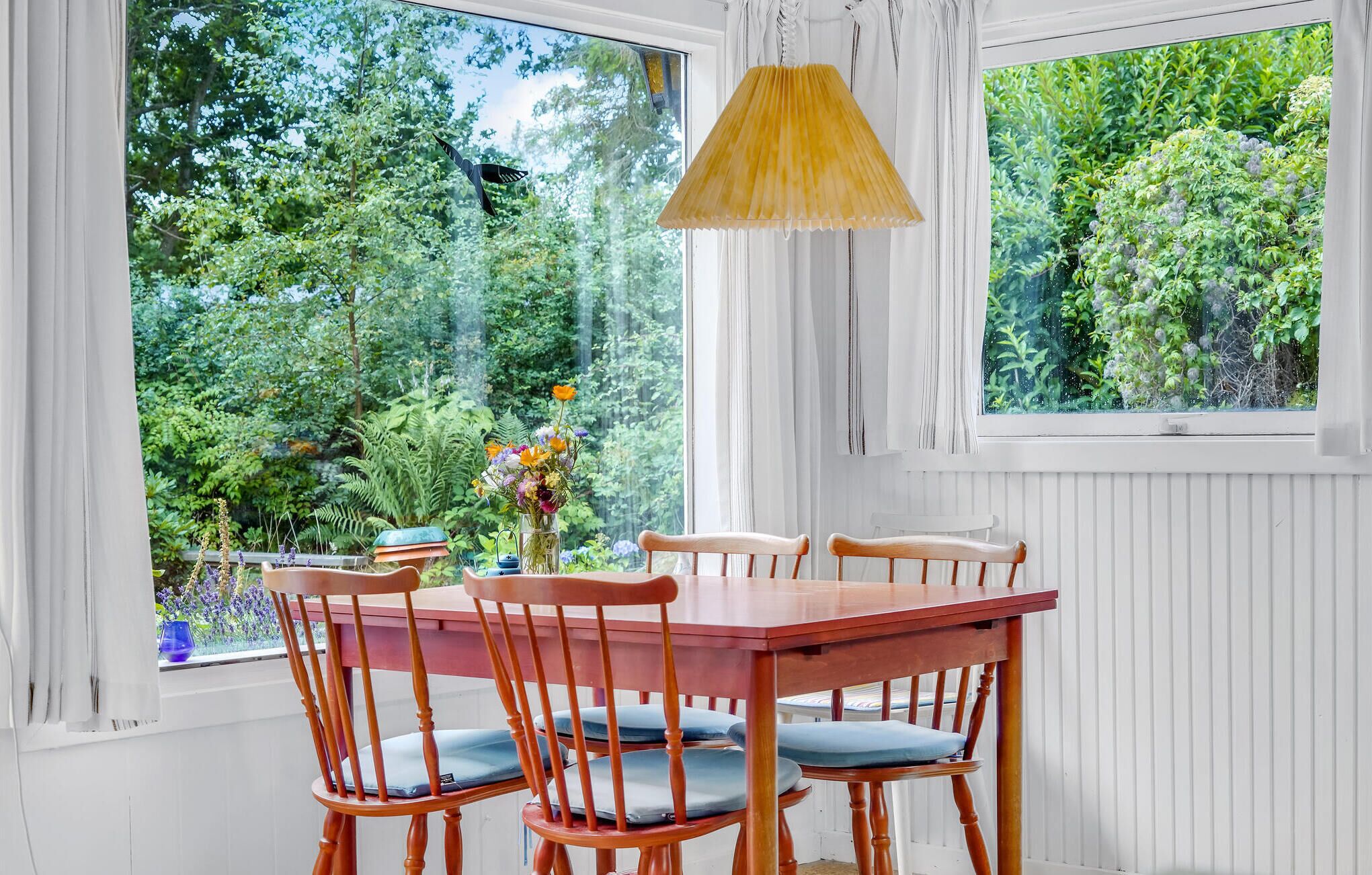 Ein Essbereich mit einem Holztisch und Stühlen, einer Blumenvase und einem Fenster mit Blick auf Grünflächen.